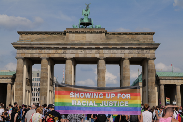Gruppe von Menschen, die vor dem Brandenburger Tor in Berlin, Deutschland, mit einer "Racial Justice"-Schleife stehen, mit den Säulen und der Statue des Tors im Hintergrund.