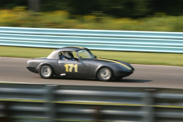 Ein Auto mit einem Helm tragenden Fahrer fährt auf einer von Barrieren gesäumten Straße, mit Bäumen im Hintergrund.