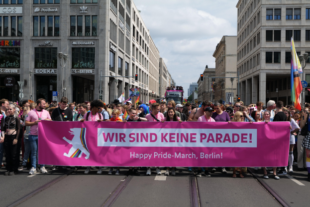 Eine Gruppe von Menschen, die eine rosafarbene Fahne mit der Aufschrift "Happy Pride March" tragen, geht eine Straße in Berlin, Deutschland, entlang. Die Straße ist von Gebäuden, Laternenmästen und Verkehrszeichen gesäumt, öber einem bewölkten Himmel.