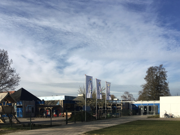 Großes Gebäude mit Metallzaun, Rasenfläche, Weg, Bäumen, Schildern, einem geparkten Fahrrad, nahen Häusern und einem bewölkten Himmel, identifiziert als neuer Spielplatz an der Schule.