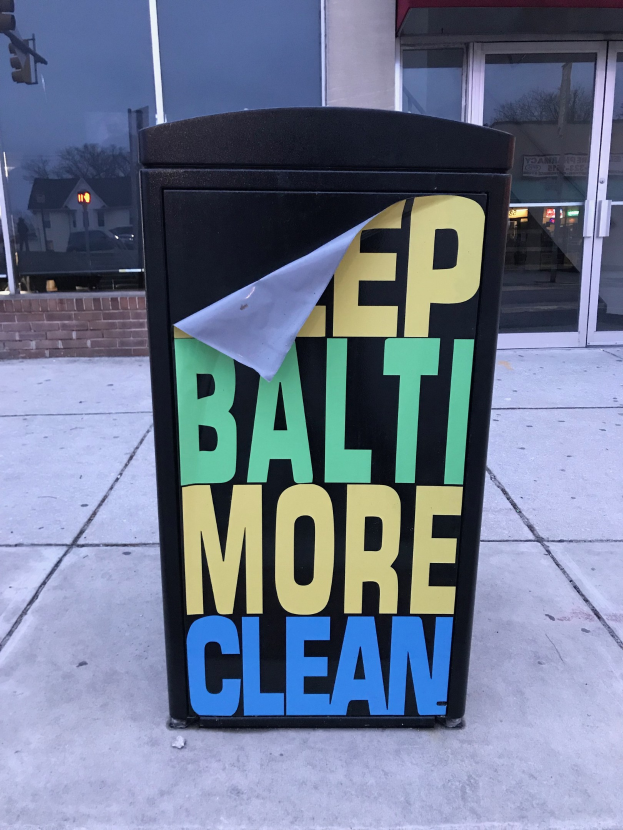 Schwarzer Mülleimer mit "Keep Baltimore More Clean"-Text auf einem Gehweg vor einem Gebäude mit Glastüren.