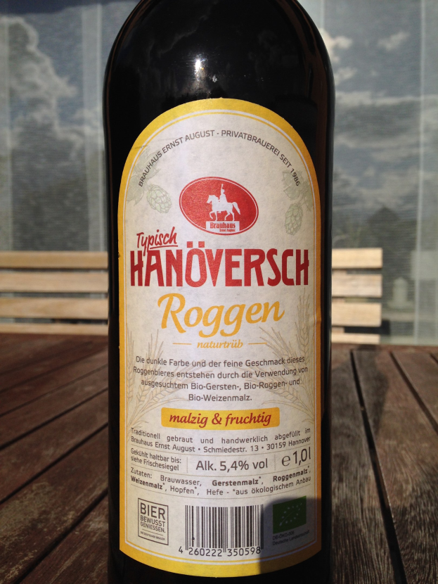 Eine Bierflasche mit Etikett steht auf einem Holztisch, im Hintergrund ist ein Glasfenster zu sehen, das ein Gebäude und den Himmel zeigt.