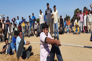 Eine Gruppe von Menschen sitzt auf einem sandigen Strand, mit einem Mann, der eine Leine in der Vordergrund hält, Bäume und Gebäude im Hintergrund und ein Tuch über einen Stuhl auf der rechten Seite, was ein Seilziehen-Wettbewerb suggeriert.