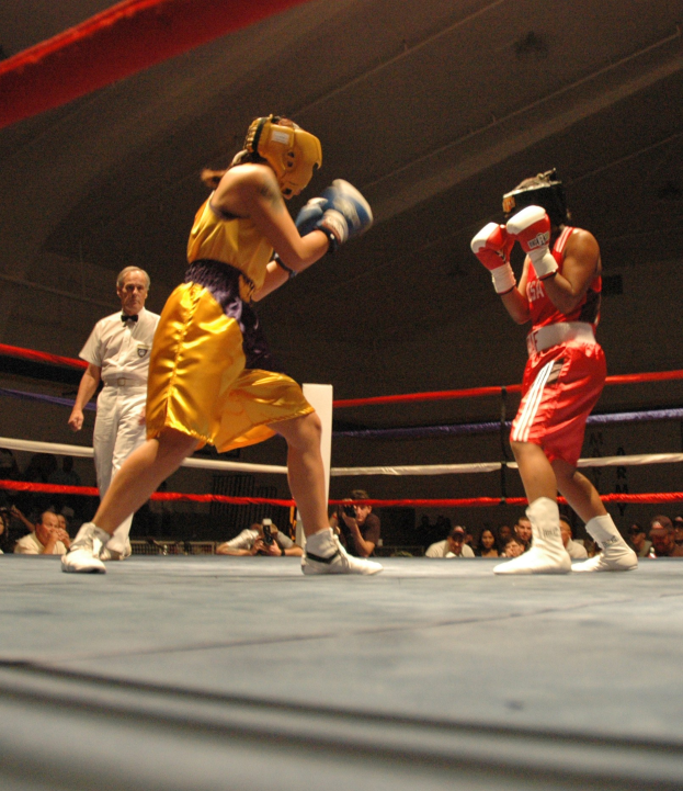 Zwei Boxer kämpfen in einem Ring, während ein Schiedsrichter beobachtet, umgeben von Fotografen und Zuschauern unter einer sichtbaren Decke.