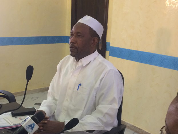 Ein Mann in einem weißen Gewand und Hut sitzt an einem Tisch mit Mikrofonen, spricht, während Papiere und Gegenstände auf dem Tisch sind, mit einer Person mit Brille rechts von ihm, in einem Raum mit einer Wand und Tür dahinter.