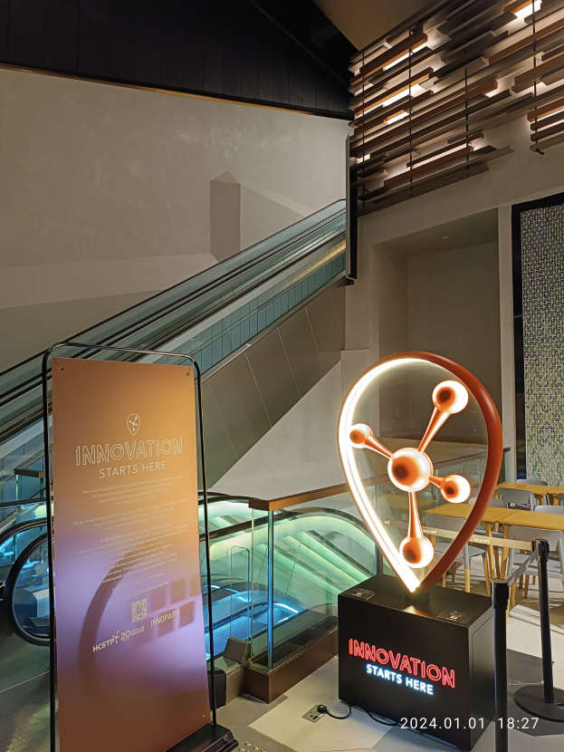 Großes Schild mit der Aufschrift "Innovation beginnt hier" in einem Gebäude mit einer Rolltreppe im Hintergrund, Tischen und Stühlen auf der rechten Seite und der HSTP20 Expo-Branding auf der linken Seite.