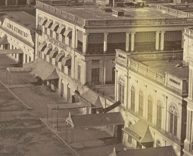 Altes Schwarz-Weiß-Foto einer Stadtstraße mit Gebäuden, Fenstern, Säulen, Namensschildern, Schuppen und Pfählen.