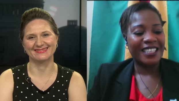 Zwei Frauen, die während eines Videoanrufs lächeln, eine in einem schwarzen und weißen Kleid, die andere in einer unbekannten Farbe, mit einer Flagge an der Wand im Hintergrund in einer öffentlichen Einrichtung.