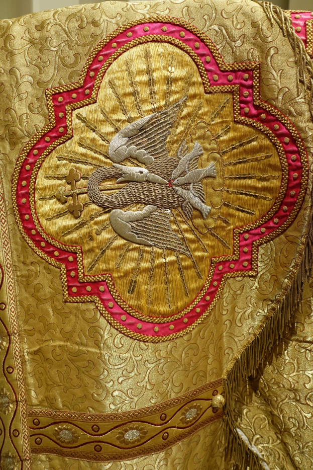 Nahaufnahme eines Priestergewandes mit aufwendiger goldener und roter Stickerei und einem luxuriösen, königlich anmutenden Design vor einer Wand im Hintergrund.
