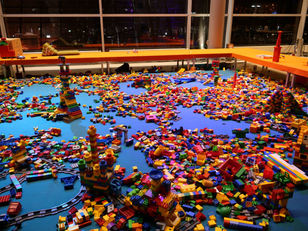 Ein großer Raum mit bunten LEGO-Baukästen, die auf dem Boden verstreut sind, umgeben von Tischen und Säulen, mit Glasfenstern im Hintergrund, die Menschen und Lichter zeigen.