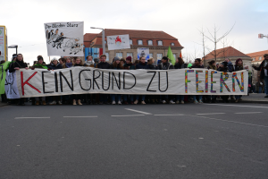 Gruppe von Menschen mit einem Transparent mit der Aufschrift "Kein Grund zu Feiern" bei einer Demonstration gegen die Sparpolitik der deutschen Regierung, im Hintergrund sind Gebäude, Bäume und ein klarer Himmel zu sehen.