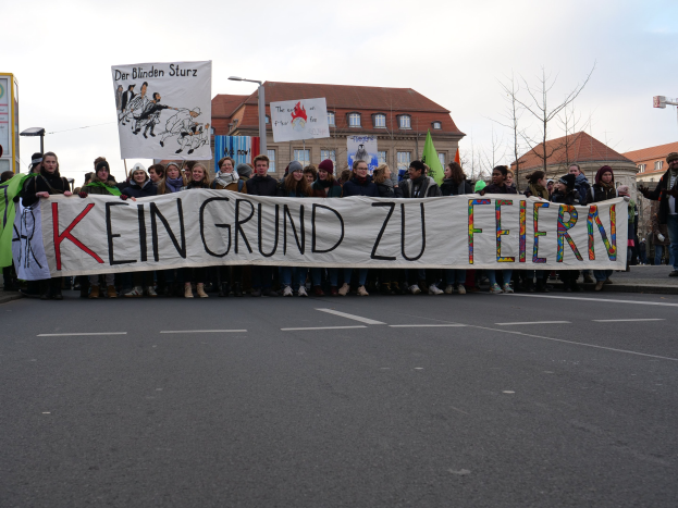 Gruppe von Menschen mit einem Transparent mit der Aufschrift "Kein Grund zu Feiern" bei einer Demonstration gegen die Sparpolitik der deutschen Regierung, im Hintergrund sind Gebäude, Bäume und ein klarer Himmel zu sehen.