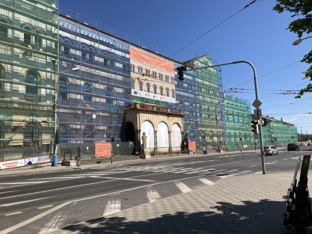 Großes Mehrfamilienhaus im Bau in Berlin, umgeben von Straßeninfrastruktur, Fahrzeugen, Fußgängern und Bäumen bei bewölktem Himmel.