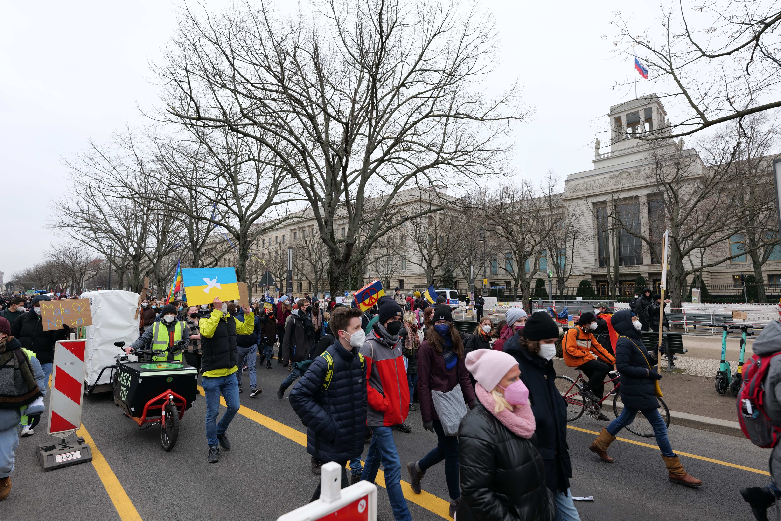 Eine große Gruppe von Menschen nimmt an einer Protestmarsch auf einer Straße teil, einige halten Schilder und andere fahren Fahrräder, mit Bäumen und einem Gebäude im Hintergrund unter einem klaren blauen Himmel.