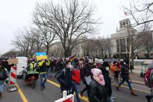 Eine große Gruppe von Menschen nimmt an einer Protestmarsch auf einer Straße teil, einige halten Schilder und andere fahren Fahrräder, mit Bäumen und einem Gebäude im Hintergrund unter einem klaren blauen Himmel.