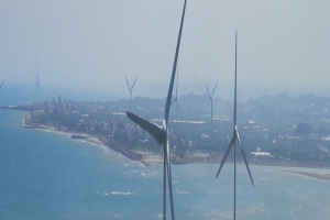 Gruppe von Windkraftanlagen im Meer in der Nähe einer Stadt, mit Wasser am Strand, Bäumen und Gebäuden im Hintergrund und einem klaren blauen Himmel.