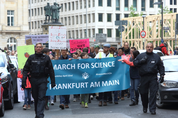 Eine Gruppe von Menschen marschiert die Straße entlang und hält ein "March for Science Frankfurt am Main"-Schild, mit Autos, Gebäuden, Statuen, Laternenmasten, Schildern und Bäumen im Hintergrund.
