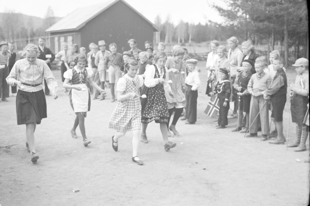 Schwarzes Foto von Kindern, die Fußball spielen, mit einer Menge Zuschauern, die Fahnen halten, Bäumen und einem Haus im Hintergrund.
