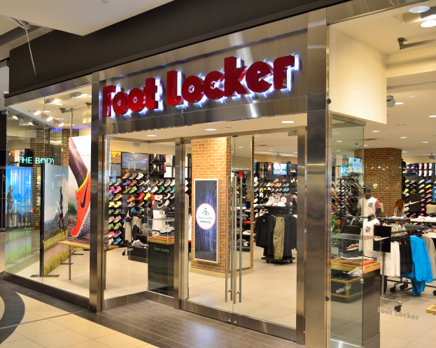 Ein Foot Locker Geschäft in einem Einkaufszentrum mit Glaswänden und -türen, Kleider an Bügeln, Schaufensterpuppen in Kleidern, Texttafeln, Deckenleuchten und andere Gegenstände.