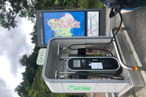 Eine Ladesäule für Elektrofahrzeuge mit einem digitalen Anzeigeboard befindet sich an der Seite einer Straße, neben einem Motorrad geparkt, und Bäume mit einem klaren blauen Himmel im Hintergrund.