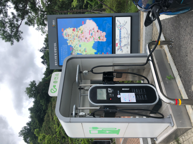 Eine Ladesäule für Elektrofahrzeuge mit einem digitalen Anzeigeboard befindet sich an der Seite einer Straße, neben einem Motorrad geparkt, und Bäume mit einem klaren blauen Himmel im Hintergrund.