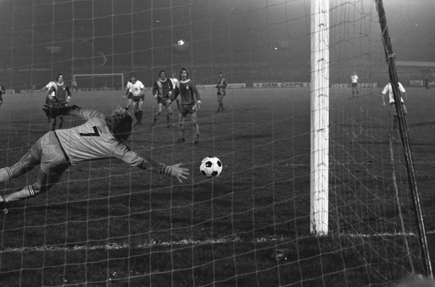 Ein Schwarz-Weiß-Foto eines Fußballspielers, der vor einem Tor einen Ball hält, umgeben von einer Gruppe von Menschen auf dem Boden. Rechts ist der Torpfosten sichtbar, im Hintergrund gibt es Werbetafeln und Lichter.