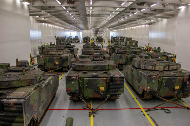 Ein grosser Militargelaenderraum mit zahlreichen Tanks in einer ordentlichen Reihe, beleuchtet von Deckenlichtern und von Waenden umgeben.