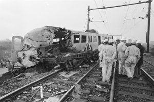 Eine Schwarz-Weiß-Szene eines Zugunfalls mit Menschen in Helmen drumherum, Strommäste, Bäume, Gebäude und einen klaren Himmel im Hintergrund.