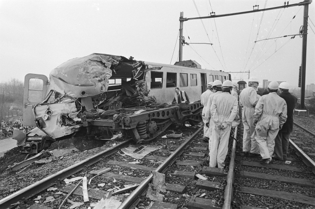 Eine Schwarz-Weiß-Szene eines Zugunfalls mit Menschen in Helmen drumherum, Strommäste, Bäume, Gebäude und einen klaren Himmel im Hintergrund.