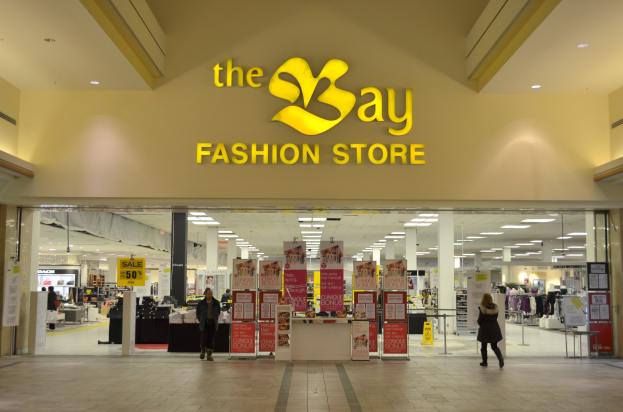 Verkaufsraum eines Bay Fashion Retail-Shops in einem Einkaufszentrum mit vorbeigehenden Kunden, Texttafeln an den Wänden, tragenden Säulen, Deckenbeleuchtung und zusätzlichen Elementen des Einkaufszentrums im Hintergrund.