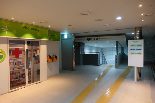 Apothekeneingang mit einem "Apotheke"-Schild auf der linken Seite, einer Tafel mit Text auf der rechten Seite, beleuchteten Lampen darüber und undeutlichen Hintergrundobjekten.