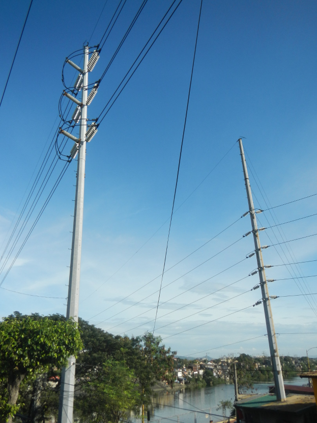 Stromleitung mit Strommasten und Kabeln, die neben einer Straße verlaufen, ein Gewässer, Bäume, Gebäude und einen klaren blauen Himmel.