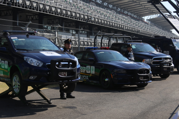 Polizeiautos vor einer Rennstrecke mit uniformierten Personen, einem Stadion im Hintergrund und Maschendrahtzaun.