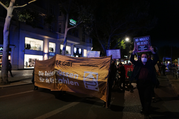 Gruppe von Menschen mit Schildern und Plakaten auf einer nächtlichen Straße in Berlin unterwegs, mit Bäumen, Gebäuden, Laternen und Fahrrädern im Hintergrund.
