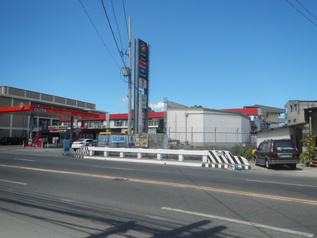 Tankstelle an einer Straßenecke mit parkenden Autos, umgeben von Gebäuden, Versorgungsmasten, Schildern, Geländern und Pflanzen, unter einem bewölkten Himmel.