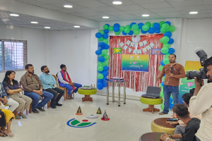 Eine Gruppe von Menschen, die auf Stühlen sitzen und einer Menge gegenübersitzen, mit einem Lautsprecher, der ein Mikrofon hält, einem Laptop auf einem Hocker, einem Tisch mit Gegenständen, einer Fahne, Ballons, einem Vorhang, einem Fenster und Deckenleuchten, was auf eine 9. Geburtstagsfeier hinweist.