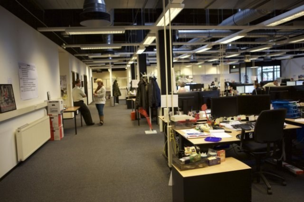Großes Großraumbüro mit Menschen, die an Schreibtischen mit Computern, Monitoren, Tastaturen und Papieren arbeiten, umgeben von Stühlen, Kisten und Büchern; Kleider hängen von Gestellen, Tafeln mit Text und Bildern sind an den Wänden angebracht und Deckenlampen erhellen den Raum, mit Glasfenstern im Hintergrund.
