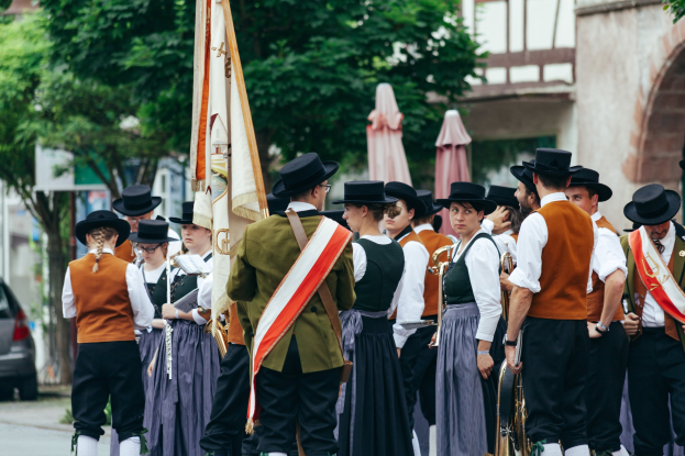 Eine Gruppe von Menschen in traditioneller bayrischer Kleidung marschiert die Straße entlang, einige halten Musikinstrumente und Fahnen, im Hintergrund sind Bäume, Gebäude und ein Auto zu sehen.