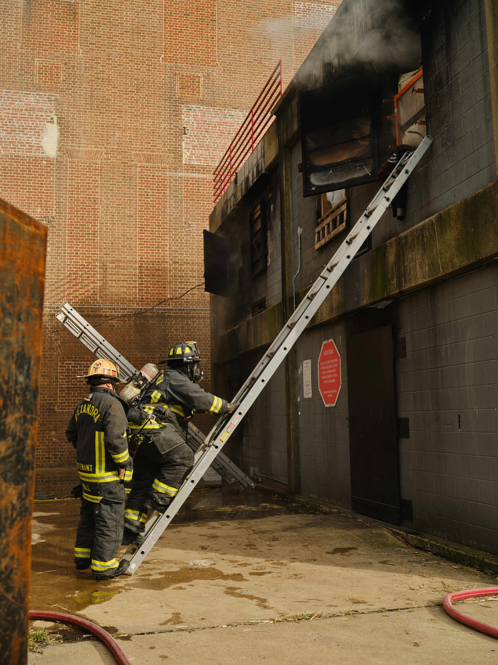 Feuerwehrleute mit Helmen klettern an einer Leiter zu einem rauchgefüllten Gebäude hoch, mit einem Metallobjekt links und Rohren an der Basis.
