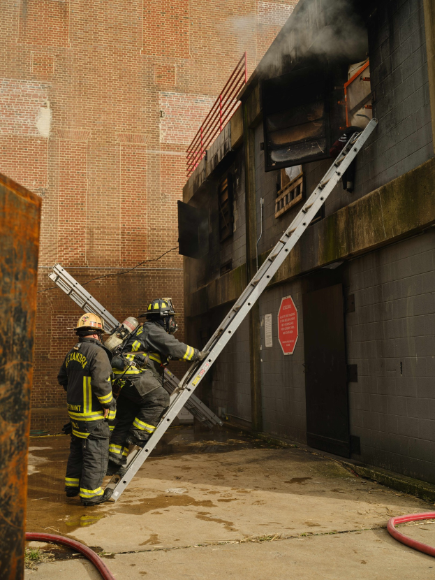 Feuerwehrleute mit Helmen klettern an einer Leiter zu einem rauchgefüllten Gebäude hoch, mit einem Metallobjekt links und Rohren an der Basis.