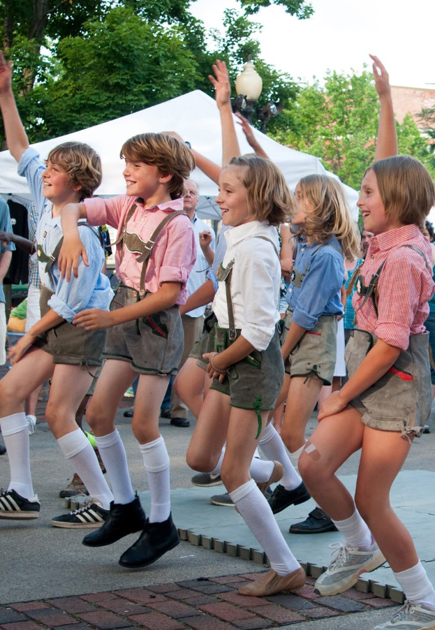 Kinder in traditioneller bayrischer Tracht, die auf einer Straße tanzen, umgeben von einer Menge, Zelten, Laternenmasten, Bäumen, Gebäuden und einem klaren blauen Himmel.
