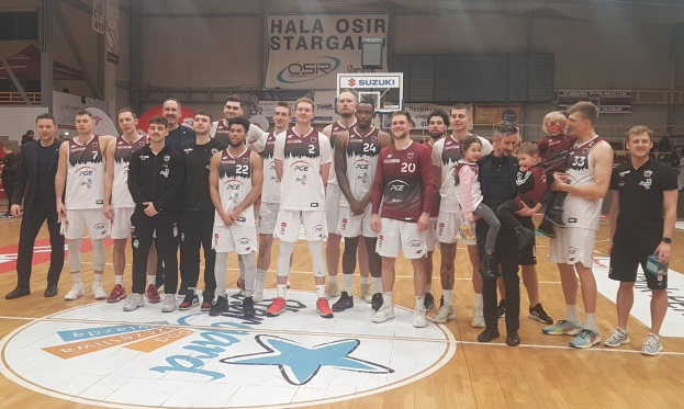 Eine Gruppe von Männern und Kindern auf einem Basketballfeld mit Schildern, die "Hala Osir Starga" lesen, sitzende Zuschauer, ein Basketballkorb und Deckenbeleuchtung.