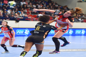 Eine Gruppe von Frauen spielt Handball auf einem Platz mit einem Ball in der Luft, während Zuschauer von Stühlen, Geländern und einer Wand im Hintergrund zuschauen.