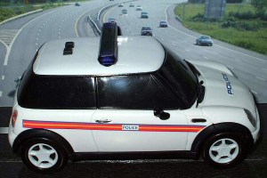 Ein weißer Polizeiwagen steht im Vordergrund mit mehreren fahrenden Autos auf der Straße dahinter, flankiert von Gras und Bäumen auf beiden Seiten in einer ländlichen Umgebung.