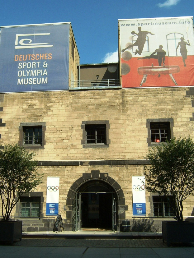 Außenansicht des Deutschen Sport & Olympia Museums in Berlin: Ein Gebäude mit Fenstern und einer Tür, umgeben von Bäumen und Bannern unter einem bewölkten Himmel.