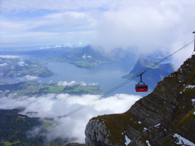Seilbahn fährt einen Berg mit einem See, Hügeln, Bäumen und Wolken im Hintergrund hoch.