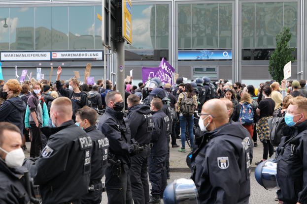 Große Gruppe von Menschen demonstriert vor einem Gebäude, einige halten Schilder und tragen Helme, mit einem Schildständer im Vordergrund und einem Baum im Hintergrund.