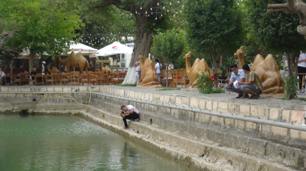 Ein Mann sitzt am Rand eines Teiches umgeben von Menschen, Bäumen, Skulpturen, Möbeln und Schirmen mit Wasser im Vordergrund und Himmel im Hintergrund.