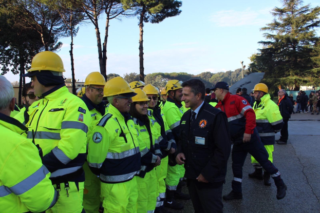 Gruppe von Einsatzkräften in gelben Westen und Helmen auf einer Straße mit Bäumen und einem klaren blauen Himmel im Hintergrund.