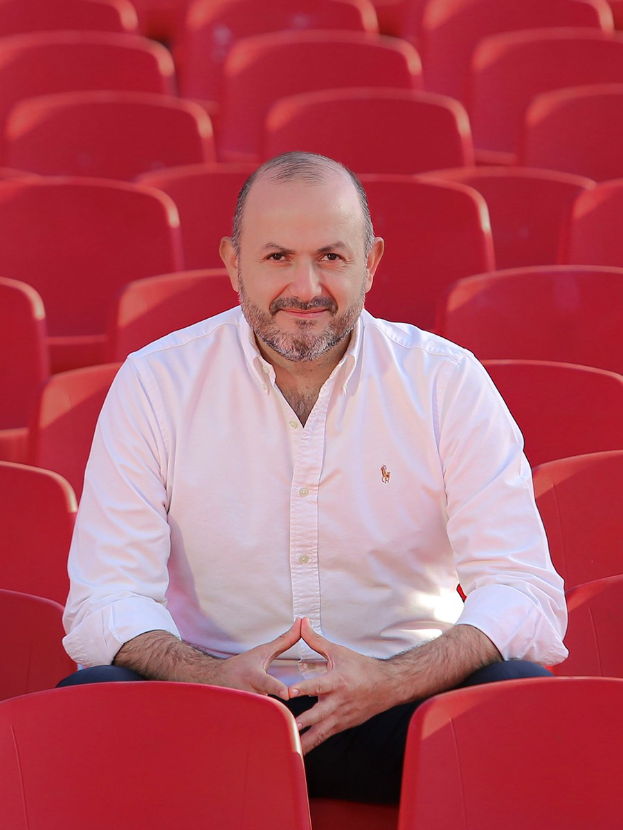 Ein Mann in einem weißen Hemd sitzt selbstbewusst in einem roten Stadionstuhl, die Hände verschränkt, mit einem ernsten Gesichtsausdruck, während Sonnenlicht im Hintergrund scheint.